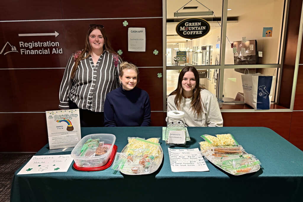 "Three young women at a St. Patrick's Day bake sale table with a green tablecloth, offering various baked goods like brownies, banana bread, and chocolate-covered pretzels. Signs on the table highlight prices, a donation jar for 'Mom's House Inc.,' and a Cash App payment option (SydneyScott24). The bake sale supports 'Mom's House Inc.' The background shows a 'Registration Financial Aid' sign and a Green Mountain Coffee shop through a glass partition."