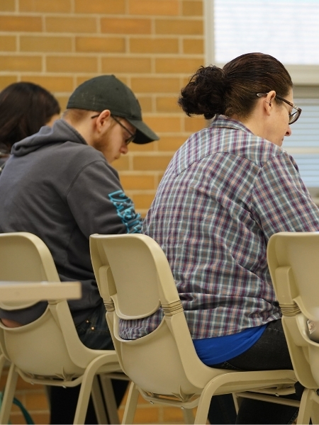 Two individuals seated in beige chairs against a tan brick wall, likely in a classroom or study setting. On the right, a woman with dark hair in a bun wears glasses and a purple, white, and grey plaid shirt over a blue undershirt. On the left, a man in a dark grey hoodie with blue text on the sleeve and a black baseball cap looks down, appearing focused. The background includes a window with horizontal blinds and overexposed light.